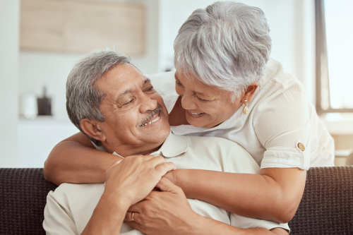 Mixed race senior couple hugging in a living room at home. Smiling elderly husband and wife holding each other and embracing in lounge. Happy playful retired man and woman bonding and feeling in love